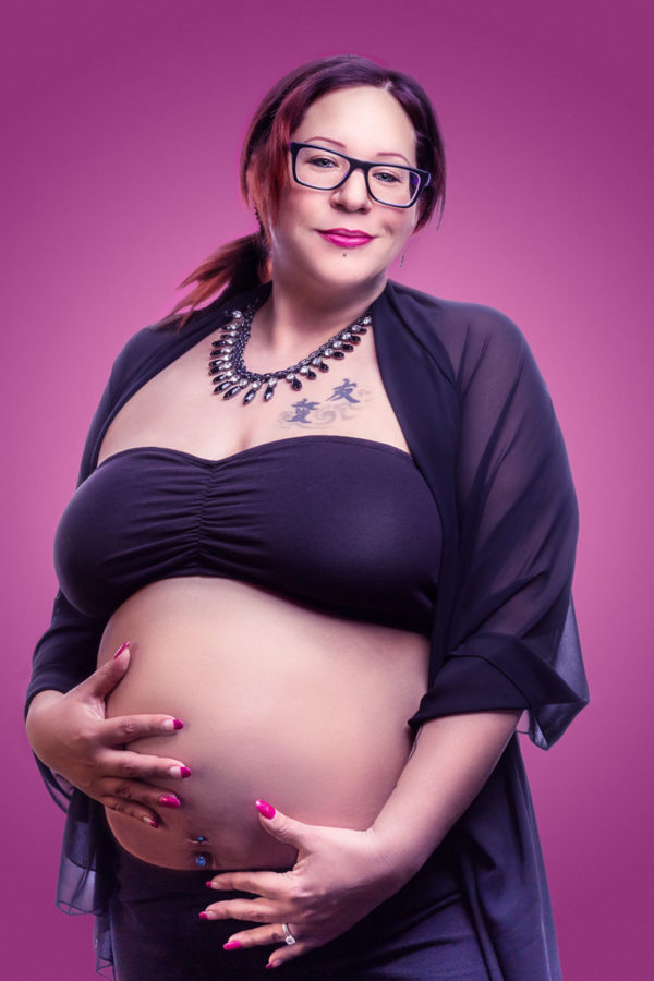 Fotograf Schwarzwald Babybauch Frau mit Brille vor rosa Hintergrund hält ihren Bauch